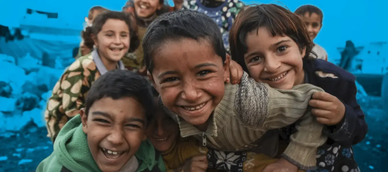 A group of smiling children hugging and laughing together in front of a blue tarp, symbolizing hope, resilience, and childhood joy despite hardship